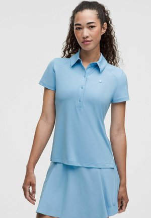 Frau mit dunklem lockigem Haar, die ein hellblaues kurzärmeliges Poloshirt und einen passenden hellblauen Rock vor neutralem Hintergrund trägt.