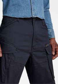 Mitten av en person som bär mörkblå cargobyxor med dragkedja och klafffickor, ihop med en ljusblå denimskjorta.