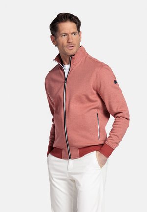 Man draagt een zalmroze jas met ritssluiting, geribde boorden en kraag, witte broek, staat met handen in de zakken en kijkt opzij.