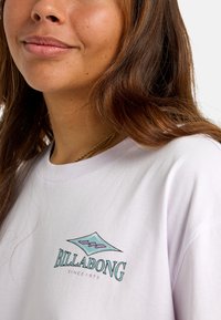 Biela bavlnená tričko s tehlovo modrým diamantovým logom a textom "BILLABONG" v čiernej farbe, s okrúhlym výstrihom a krátkymi rukávmi.