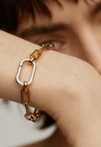 Pulsera de cadena metálica de oro y plata con un diseño de eslabones sobredimensionados. Textura suave, con un detalle de cierre rectangular.