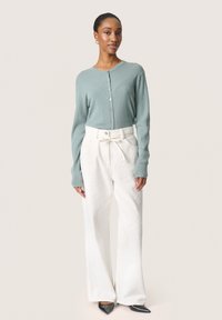 Cardigan a coste verde chiaro con bottoni frontali abbinato a pantaloni bianchi a gamba larga con texture. Il modello indossa scarpe nere a punta.