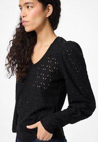 Svart bluse i blonder med V-hals, puffede ermer og intrikate utskjæringer med bladmønster. Teksturert stoff gir dybde og dimensjon.