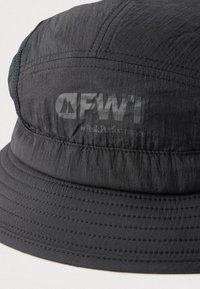 Chapeau bob noir en tissu léger avec bords cousus et logo embossé sur le devant. Texture lisse et forme souple.