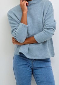 Osoba v světle modrém svetru a modrých džínách se zkříženýma rukama, jednou rukou se dotýkající brady, stojící u bílé zdi.