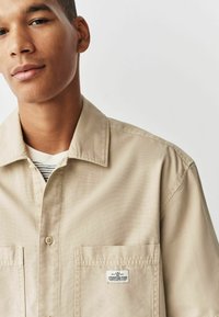 Chemise beige à boutons avec un col pointu, deux poches avant et une étiquette sur la poche gauche. Tissu lisse et léger.