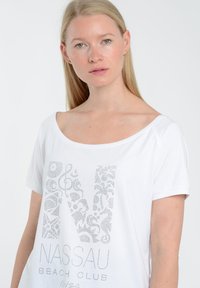 Hvid t-shirt med korte ærmer og bred udskæring. Forsiden har et sølvfarvet dekorativt print af bogstavet "N" og teksten "NASSAU BEACH CLUB."