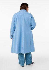 Svetlo modra jakna iz gladkega materiala z žepkim ovratnikom, dolgimi rokavi in hrbtnim razporkom, v kombinaciji z širokimi kavbojkami.