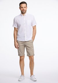 Valkoinen lyhythihainen pellavapaita, jossa napitus edessä ja vasemmassa rintataskussa, yhdistettynä käärittyihin beigekäisiin shortsit ja valkoisiin tennareihin. Yksinkertainen muotoilu.