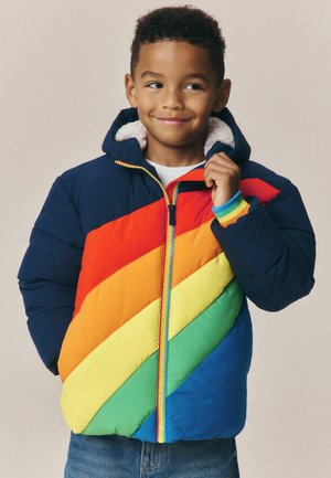 Niño pequeño que lleva una chaqueta acolchada a rayas de colores del arcoíris con capucha y vaqueros azules, sonriendo y sujetando el cuello de la chaqueta.