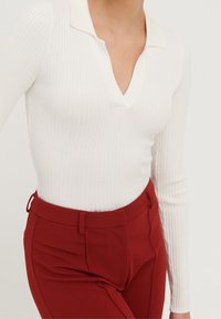 Femme portant un haut à manches longues côtelé blanc avec un col en V et un pantalon rouge taille haute ajusté.