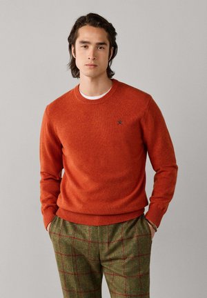 Joven con cabello oscuro que lleva un suéter naranja y pantalones verdes a cuadros, de pie con las manos en los bolsillos frente a un fondo gris.
