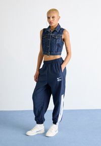 Persoon met kort blond haar, gekleed in een mouwloos denimvest en marineblauwe trainingsbroek met witte strepen, staande op een blauwe vloer tegen een witte muur.