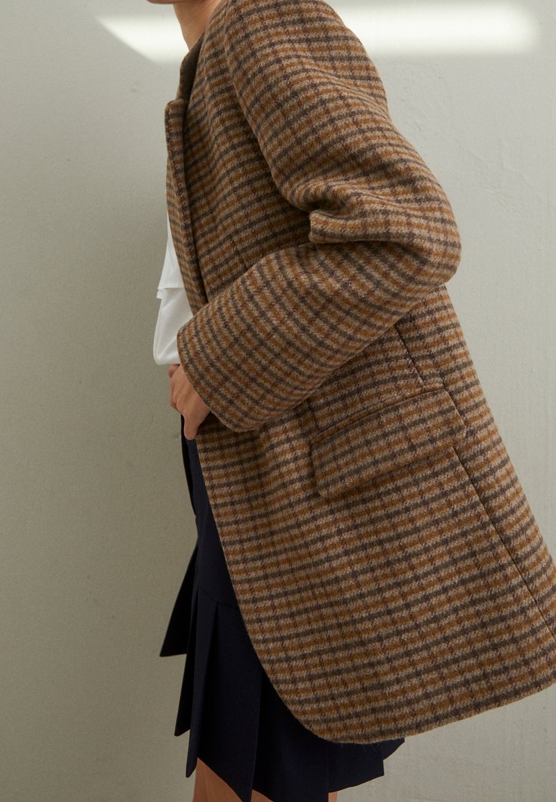 Personne portant un manteau à carreaux marron et beige sur un haut blanc et une jupe noire, debout contre un mur clair uni.