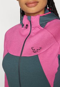 Pink and dark teal zip-up jacket with a hood. Features a textured fabric and a black logo on the chest, with contrasting color panels.