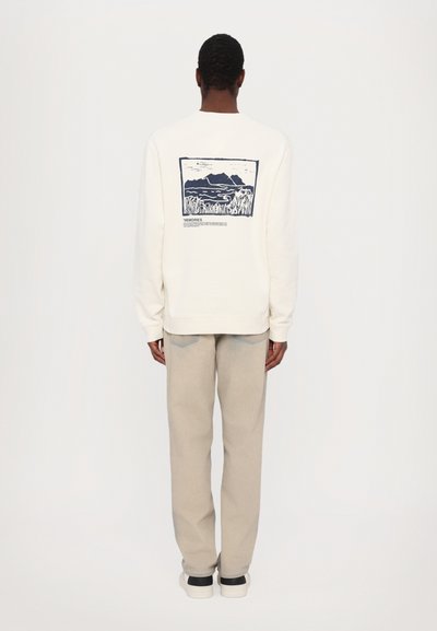 Sweatshirt crem cu un imprimeu grafic de munți și apă în nuanță de navy, având mâneci lungi și o croială lejeră. Purtat cu pantaloni de culori deschise.