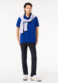 Homme portant un polo bleu avec un pull mauve clair sur les épaules, un jean foncé et des baskets blanches, debout sur fond blanc.