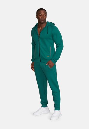 Mężczyzna stojący, ubrany w ciemnozieloną bluzę rozpinaną na zamek i dopasowane dresy oraz białe sneakersy, ręce w kieszeniach, patrzy w lewo.