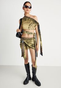 Frau, die ein gemustertes gelb-grünes, gebundenes Crop-Top und einen Minirock trägt, schwarze knielange Stiefel, dunkle Sonnenbrillen, Creolen und eine schwarze Handtasche dabei hat.