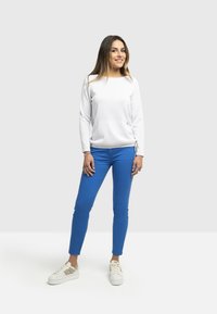 Top bianco a maniche lunghe con dettaglio arricciato sul lato, abbinato a pantaloni attillati blu brillante e sneakers bianche con accenti dorati.