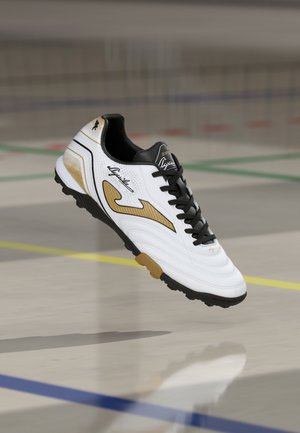 Scarpa da calcio bianca con accenti dorati e neri, materiale texturizzato, design con lacci e suola in gomma con tacchetti multidirezionali.