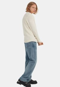 Witte, gestructureerde gebreide trui, blauwe denim spijkerbroek en zwarte schoenen. De trui heeft een geribde onderrand en mouwen, de spijkerbroek heeft een losse pasvorm.