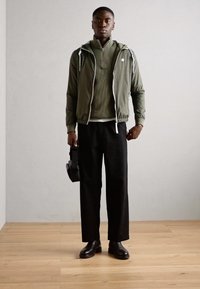 Olivgrüne leichte Jacke mit weißen Kordeln, über einem strukturierten dunklen Oberteil getragen, kombiniert mit lockeren schwarzen Hosen und schwarzen Stiefeln.