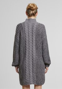 Femme aux cheveux blonds en chignon, portant une robe en tricot à grosse maille grise, vue de dos sur un fond uni.