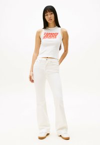 Top de tirantes blanco con logo rojo "TOMMY JEANS NEW YORK CITY", combinado con jeans acampanados blancos y zapatillas blancas con suelas marrones.