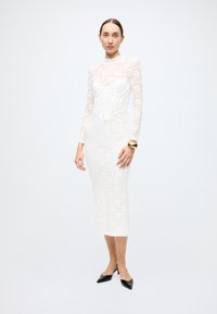 White lace dress with long sleeves, featuring a fitted bodice and floral pattern. The dress has a midi length and a smooth texture. Black pointed shoes.