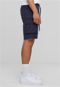 Marinefarbene Cargo-Shorts mit zwei großen seitlichen Taschen und schwarzem Kordelzug. Getragen mit weißen Sneakers und Socken in Crew-Länge.