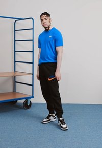 Maglietta Nike blu, pantaloni della tuta neri con logo arancione e sneakers bianco-nero. In piedi accanto a un carrello metallico blu su un pavimento blu.