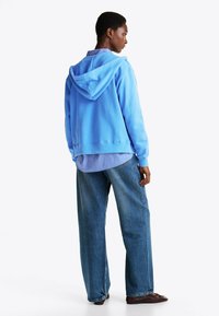 Sudadera azul claro hecha de un tejido suave, con capucha, llevada sobre una camiseta a rayas azules y jeans de mezclilla holgados.