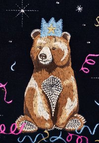 Broderad björn i nyanser av brunt och kräm, bär en blå krona med en gul stjärna. Svart bakgrund med färgglada virvlar och stjärnor.