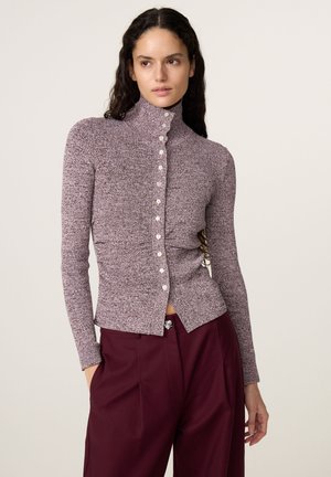 Vrouw met lang krullend haar, gekleed in een aansluitende paarse trui met knopen en hooggetailleerde donkerrode broek, staand tegen een effen achtergrond.