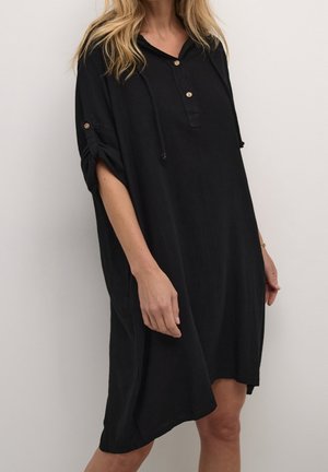 Kvinde iført løs, sort knælængde kjole med knaplukket krave og opkrogede ærmer mod ensfarvet baggrund.