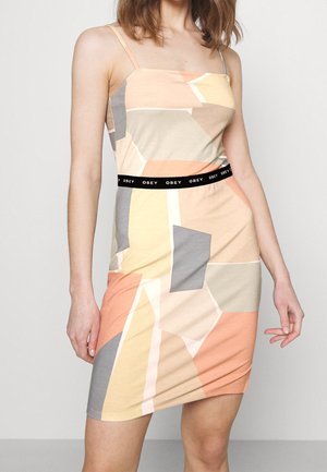 Kobieta w sukience bez rękawów z geometrycznym wzorem w kolorach brzoskwiniowym, beżowym, szarym i kremowym oraz z czarnym paskiem z napisem "OBEY".