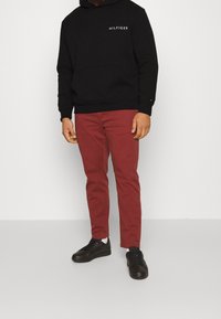 Svart huvtröja med "HILFIGER"-logotyp, röda byxor och svarta skor. Huvtröjan har en känguruficka; byxorna är slim-fit med en slät textur.