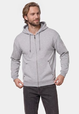 Mand iført lys grå lynlåshoodie med lommer og snore, holder i hoodieens kant, mod en ensfarvet lys baggrund.