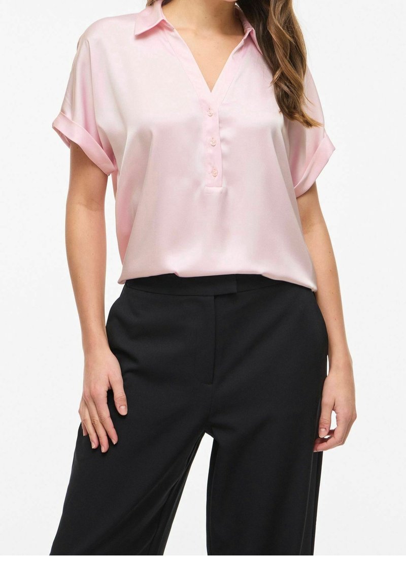 Femme portant un chemisier rose pâle à manches courtes boutonné, rentré dans un pantalon noir taille haute, visible du cou aux hanches.
