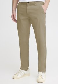 Beige cotton chinos with flat front, side pockets, and belt loops, paired with white sneakers. Smooth texture and tailored fit.