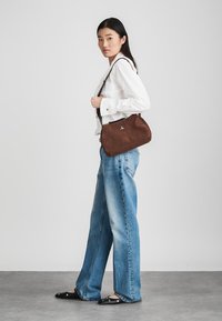 Ruda suedinė petinė rankinė su kreiva forma ir sidabriniais akcentais, nešiojama asmens, vilkinčio baltą marškinį ir mėlynus džinsus, prieš šviesų foną.