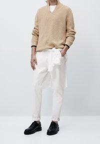 Beżowy dzianinowy pulower z głębokim dekoltem w serek, białe spodnie z podwiniętymi nogawkami oraz czarne buty na sznurowadła, stojące na neutralnym tle.