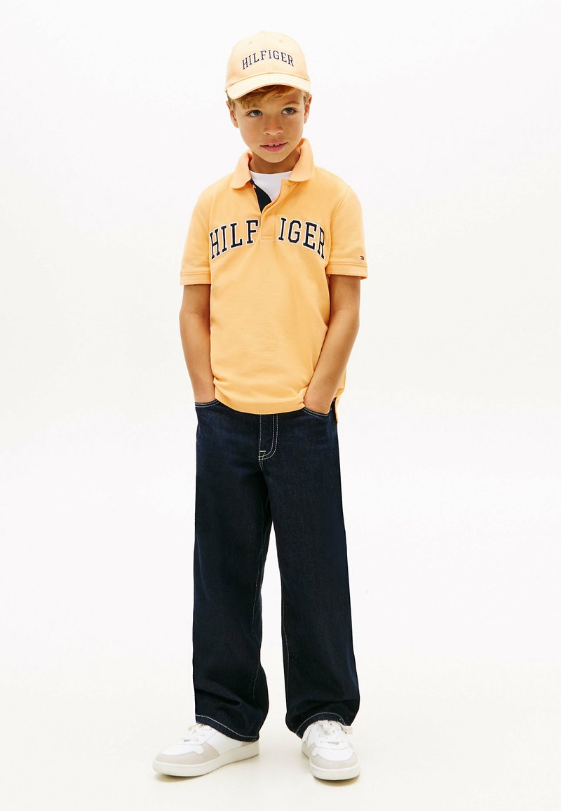 Jeune garçon portant une casquette jaune "Hilfiger" et un polo, un jean large foncé et des baskets blanches, debout les mains dans les poches.