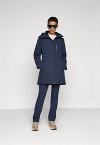 Mörkblå vattentät parka med justerbar huva, framficka med dragkedja och sidofickor; i kombination med mörkblå byxor och beigefärgade sneakers.