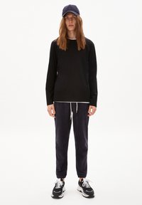 Schwarzes Langarmshirt, marinblauer Hut, dunkelgraue Jogginghosen mit weißen Kordeln und schwarze Sportschuhe mit weißen Akzenten, stehend vor einem schlichten Hintergrund.