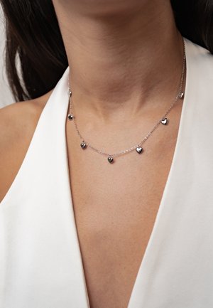 Close-up of a person wearing a silver necklace with seven small heart-shaped pendants on a silver chain and a white sleeveless top.