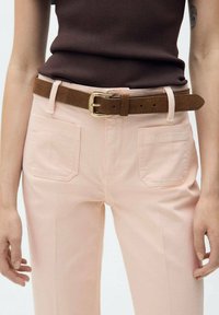 Pantalons ajustés rose clair avec poches avant et arrière, assortis à une ceinture en suède marron avec une boucle dorée. Texture de tissu lisse.