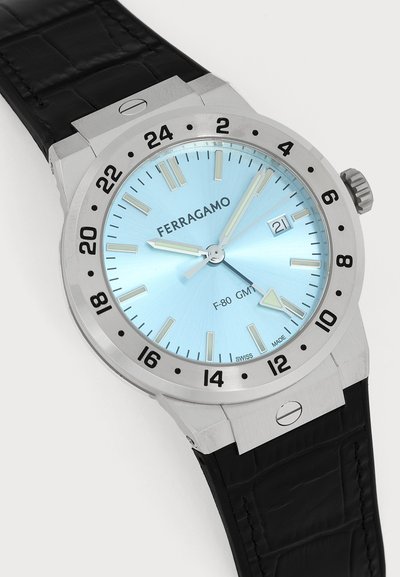 Montre-bracelet Ferragamo F-80 GMT argentée avec cadran bleu clair, lunette noire 24 heures, bracelet en cuir noir et affichage de la date à trois heures.
