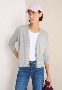 Cardigan grigio chiaro con scollatura a V e bottoni frontali, abbinato a una t-shirt bianca e jeans blu. Tiene un oggetto bordeaux con una mano.
